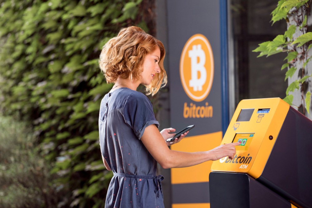 blond woman with smart phone buying bitcoin from bitcoin atm machine 