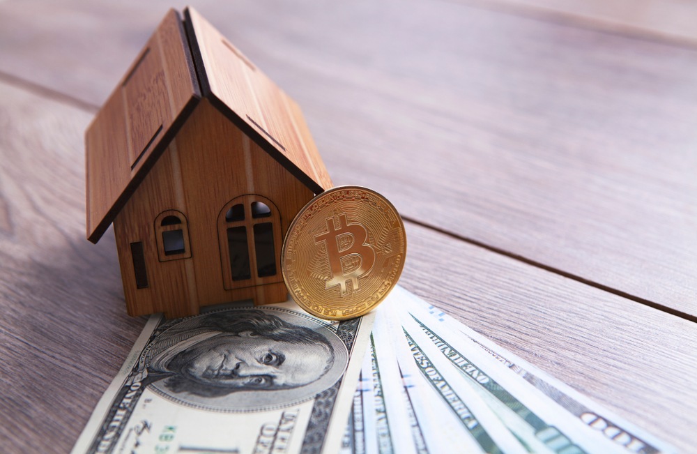 A small wooden house model sitting on stop of hundred dollar american bills and a physical bitcoin 