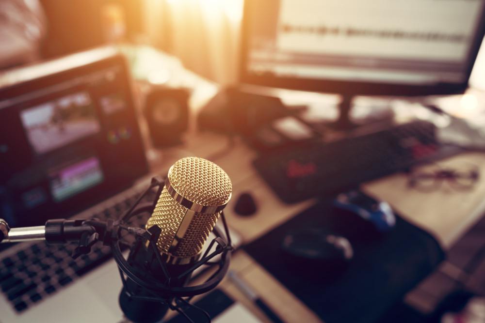 An in-focus microphone above a blurred out computer, monitor, laptop, and various related tools. 