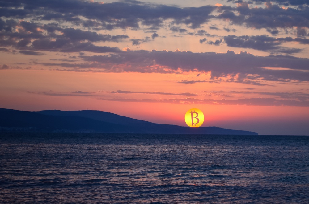 gold colored bitcoin logo in place of a rising sun over the mountains and ocean in the forefront 