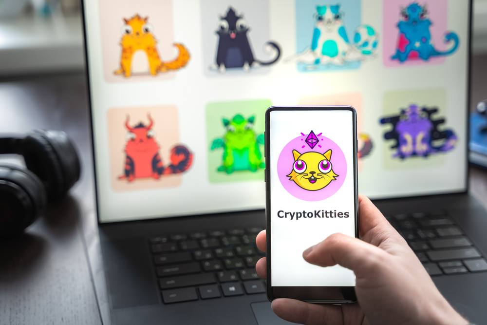 Cryptokitties logo on a mobile phone with a variety of blockchain based kitties in the background on a laptop 