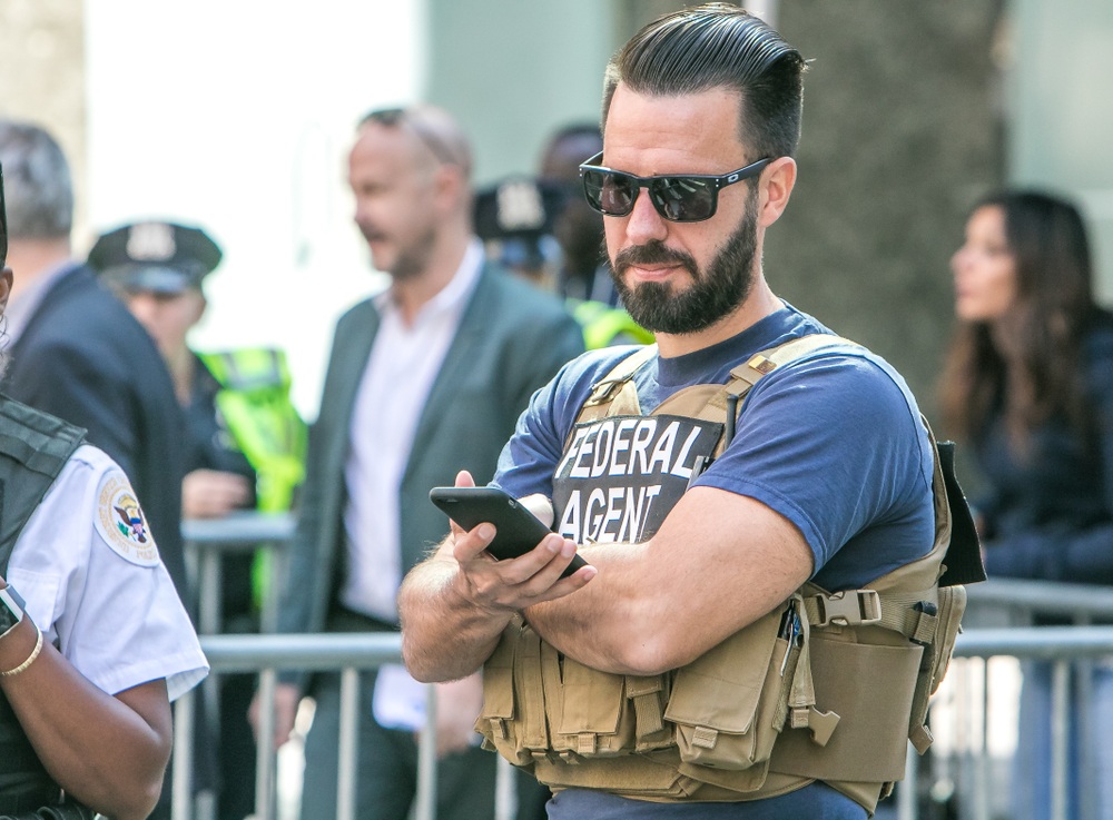 A man wearing a Federal Agent vest, with slicked back hair and glasses, with out of focus people going through a screening station in the background.  