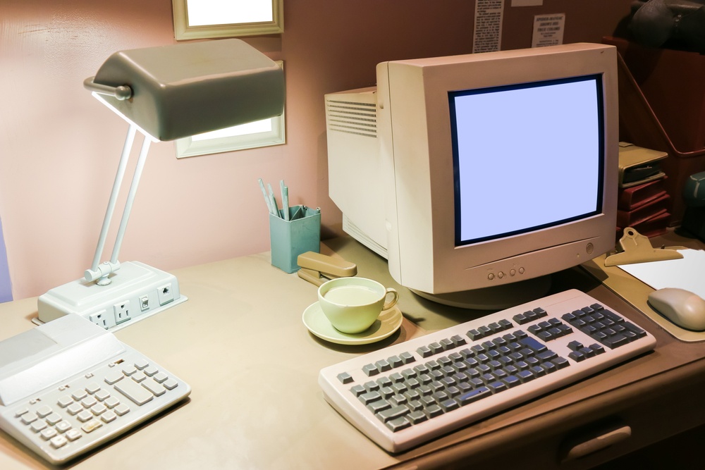 An old school computer with a keyboard on a desk with a lap, calculator, cup of milk, and more 