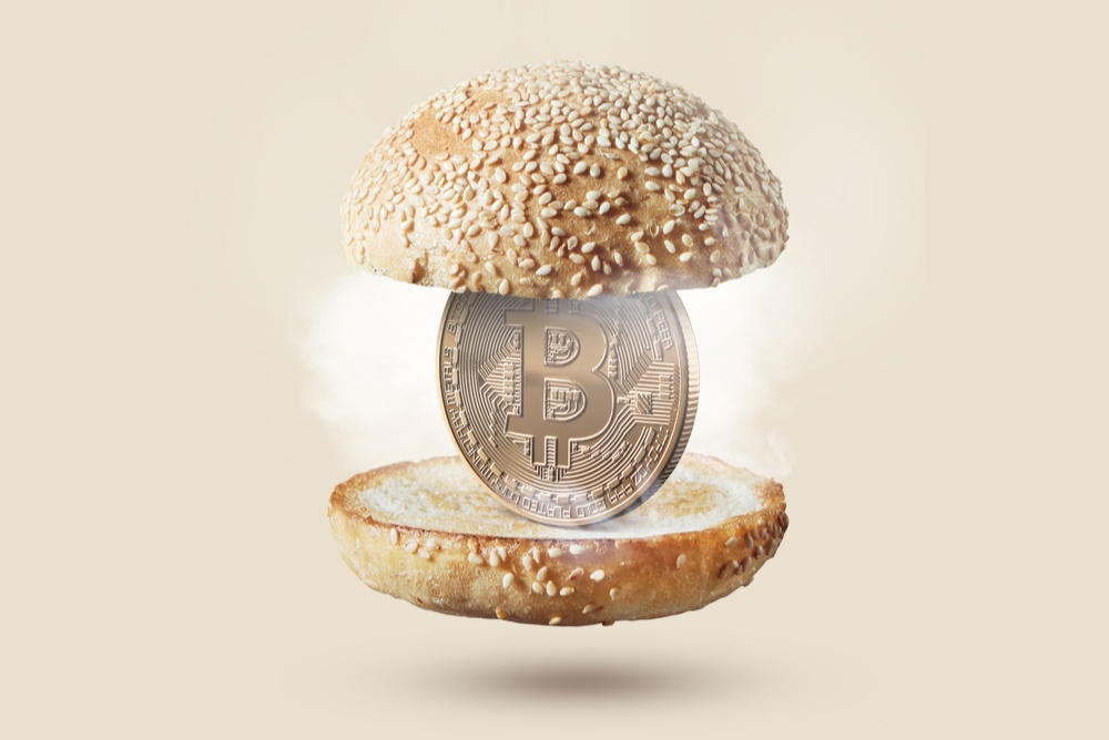 A physical bitcoin standing upright between two buns of a burger, against a beige background.  