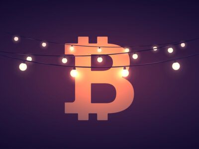 A bitcoin logo against a dark background, with strings of lights in front of it, showcasing the idea of celebrating bitcoin or bitcoin holidays&nbsp;