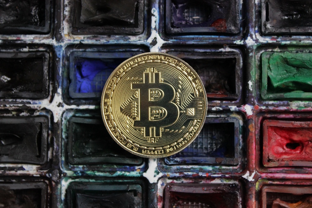 A paint palate of different colors, where many of the paints have been mostly used, with a physical bitcoin coin on top.  