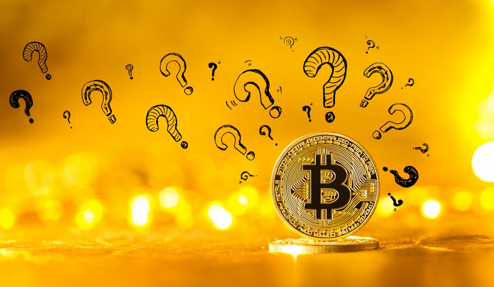 A physical bitcoin lying down and another one standing on top of it, against a yellow background, surrounded by many question marks 