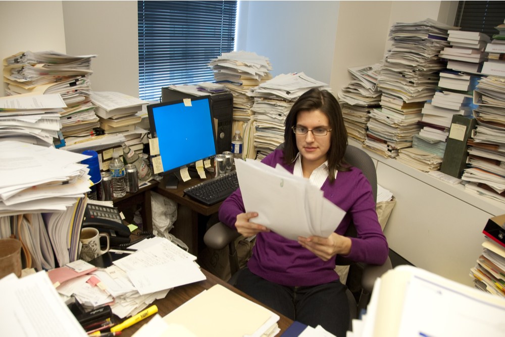 government bureaucrat reading papers with thick stacks of paper in the background. understanding and following complex laws requires resources, and can slow down growth & innovation 