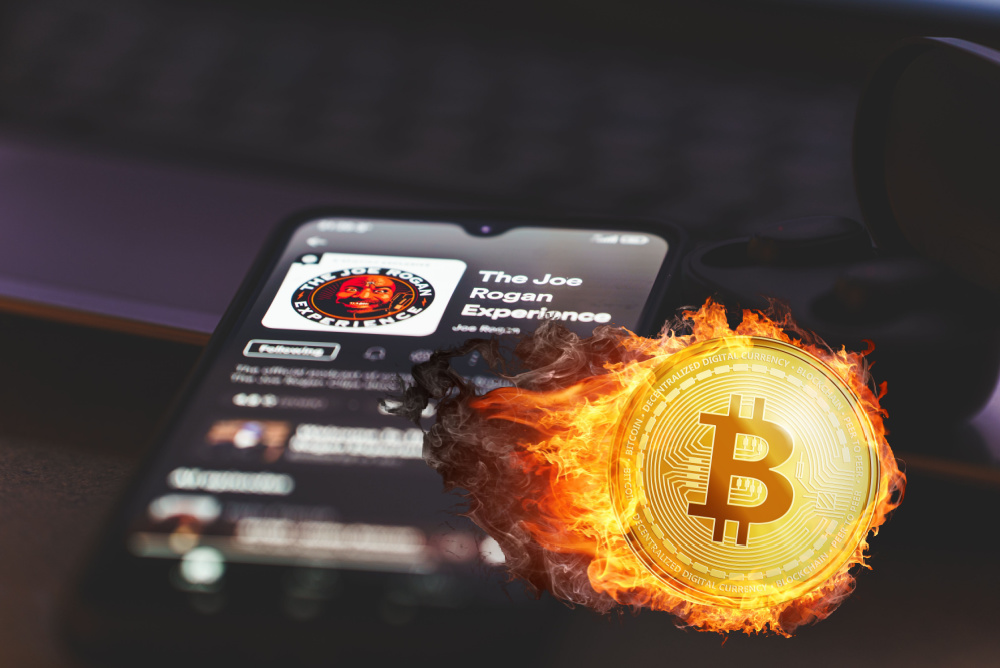 joe rogan on bitcoin. joe rogan podcast playing on smartphone with bitcoin logo on fire in foreground 