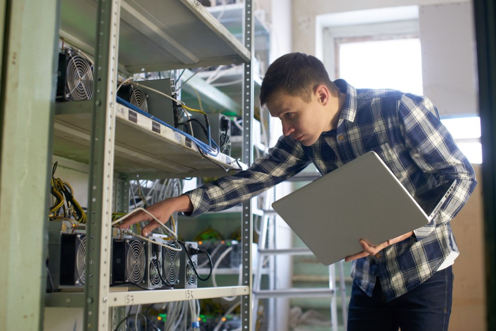 man inspecting a small rack of bitcoin ASICs with a laptop computer 